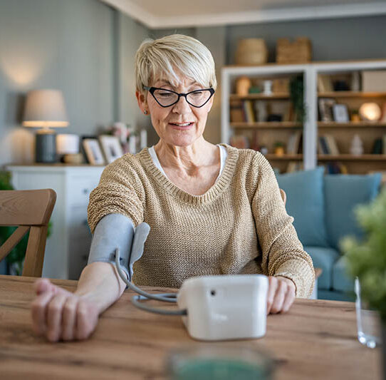 Eine Frau mit kurzen, grauen Haaren sitzt in ihrem Esszimmer und misst ihren Blutdruck mit einem Gerät mit Oberarmmanschette.