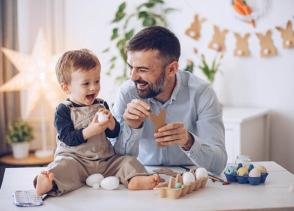 DIY zu Ostern: Deko und Geschenke Basteln: Ein Mann hat einen Papphasen und einen Stift in der Hand. Auf dem Tisch sitzt ein Kleinkind, es hält ein Ei in der Hand. Vater und Kind lachen.