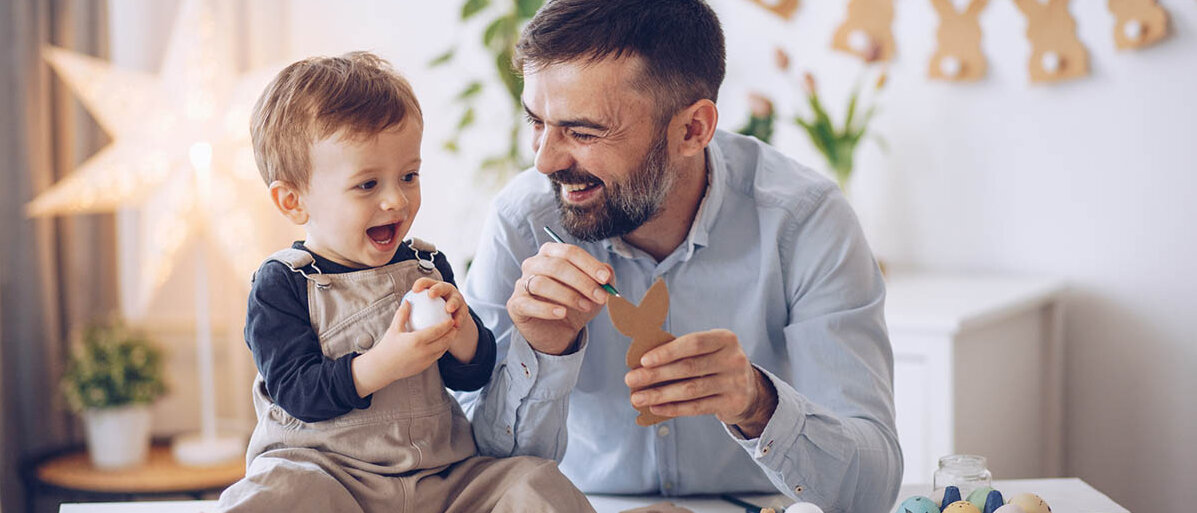 Basteln: Ein Mann hat einen Papphasen und einen Stift in der Hand. Auf dem Tisch sitzt ein Kleinkind, es hält ein Ei in der Hand. Vater und Kind lachen.