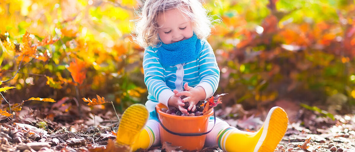 Ein kleines Kind sitzt im Herbst auf dem Boden und spielt mit einem Eimer voller Kastanien.