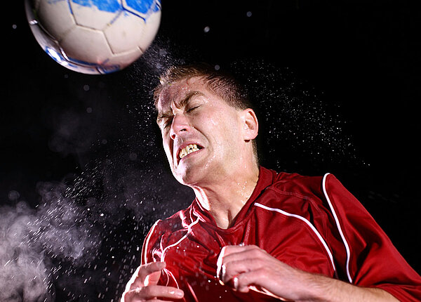 Ein Fußballer in rotem Trikot hat gerade einen Kopfball gespielt. Der Ball ist noch nah an seinem Gesicht, er hat die Arme an den Körper gezogen, verzieht das Gesicht und um seinen Kopf herum fliegen Schweißtropfen.
