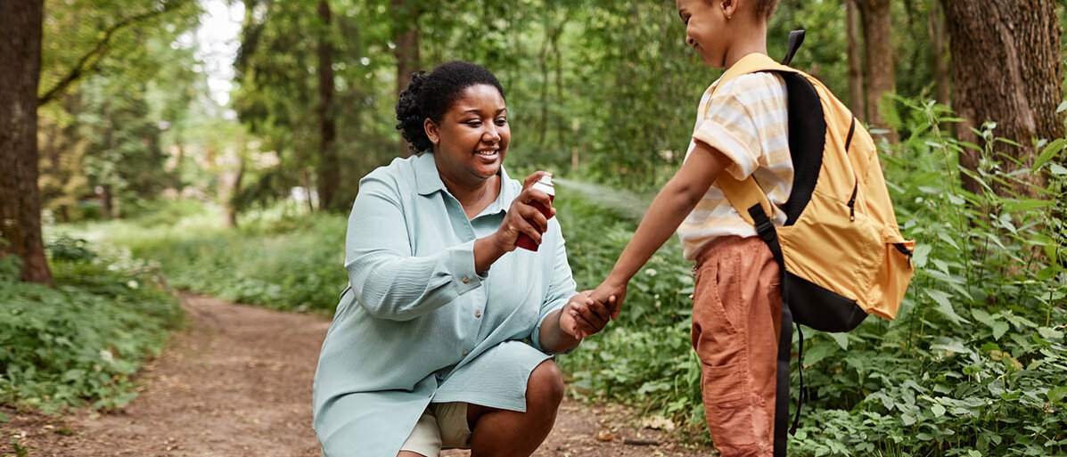 Insektenstichen wie MĂĽckenstichen vorbeugen und sie behandeln Mutter und Kind im Wald: Die Mutter kniet sich hin und sprĂĽht ihrer Tochter ein Spray auf den Arm