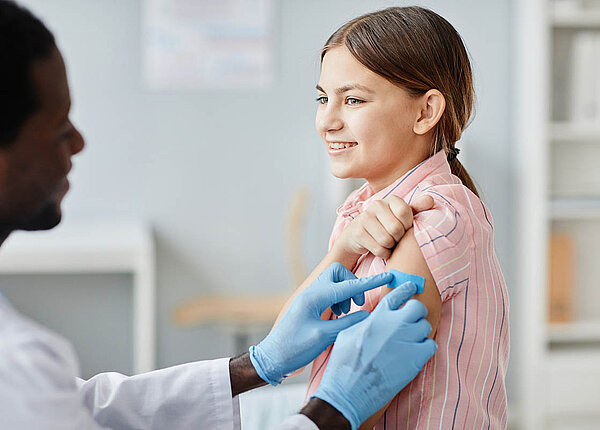 Neuer Coronaimpfstoff: Der COVID-19-Impfstoff Nuvaxovid® JN.1 Ein Arzt klebt einer Teenagerin ein Pflaster auf den Oberarm.