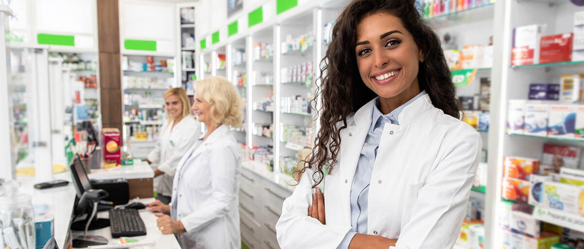 Eine junge Frau in weißem Kittel steht in der Apotheke hinter dem HV-Tisch. Sie hat die Arme vor dem Oberkörper verschränkt und lächelt selbstbewusst. Im Hintergrund stehen zwei Kolleginnen und blicken Richtung Kundschaft.