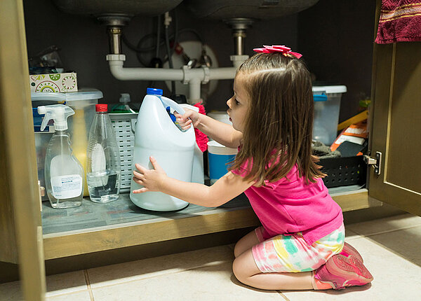 Kleines Mädchen sitzt auf dem Boden vor einem geöffneten Schrank und hat einen Reiniger in der Hand.