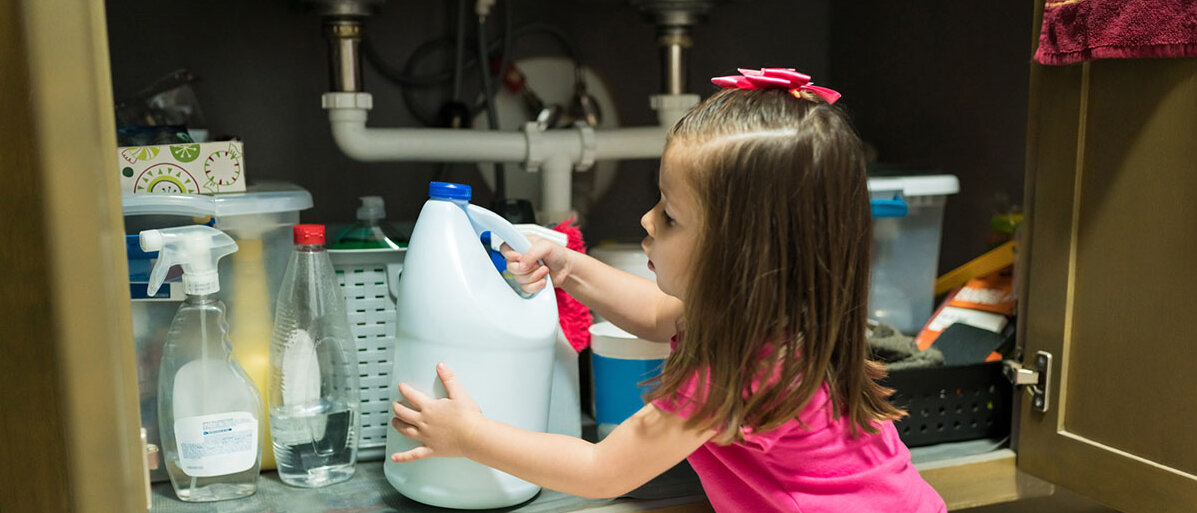 Kleines Mädchen sitzt auf dem Boden vor einem geöffneten Schrank und hat einen Reiniger in der Hand.