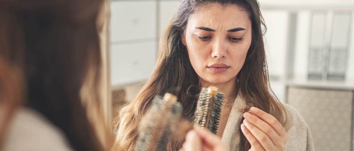 Eine Frau steht vor dem Spiegel und zieht besorgt ein Büschel Haare aus ihrer Haarbürste.