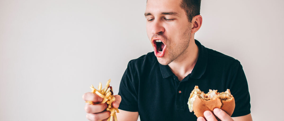 Ein Mann hält in der rechten Hand viele Pommes frites, in der linken einen angebissenen Burger. Mit gierigem Blick beißt er in die Fritten.