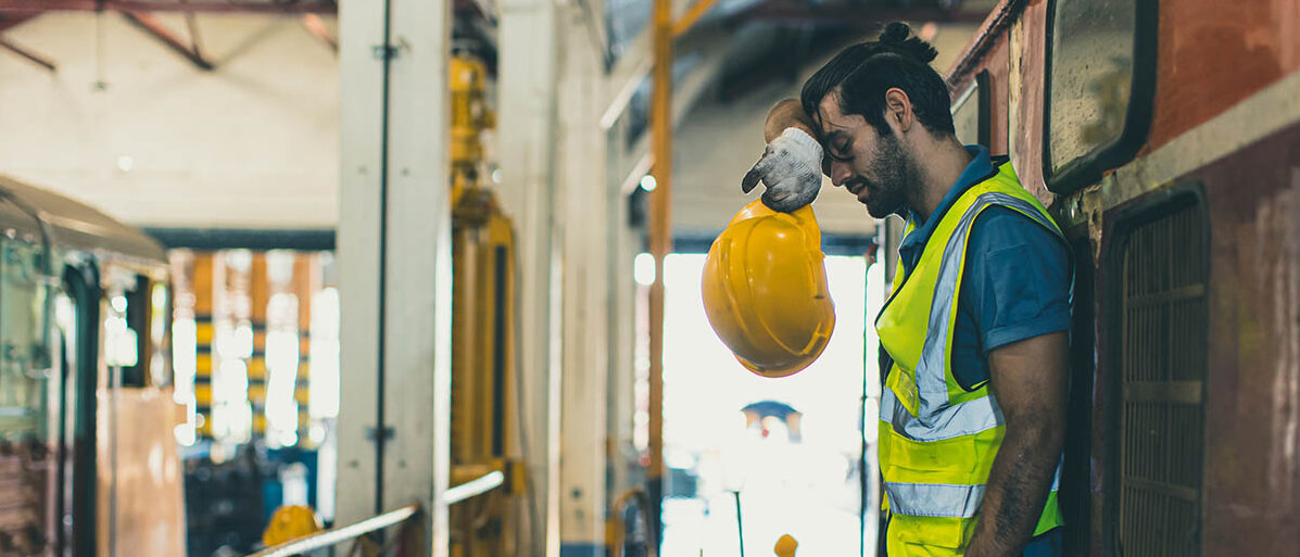 Was ist die richtige maximale Arbeitszeit pro Tag, wie viele Pausen brauchen wir, wie flexibel können wir in Zukunft arbeiten? Ein Zugmechaniker in dreckiger Arbeitskleidung lehnt erschöpft mit dem Rücken an einem Zug, hat den Helm abgenommen und wischt sich mit dem Handrücken Schweiß von der Stirn.