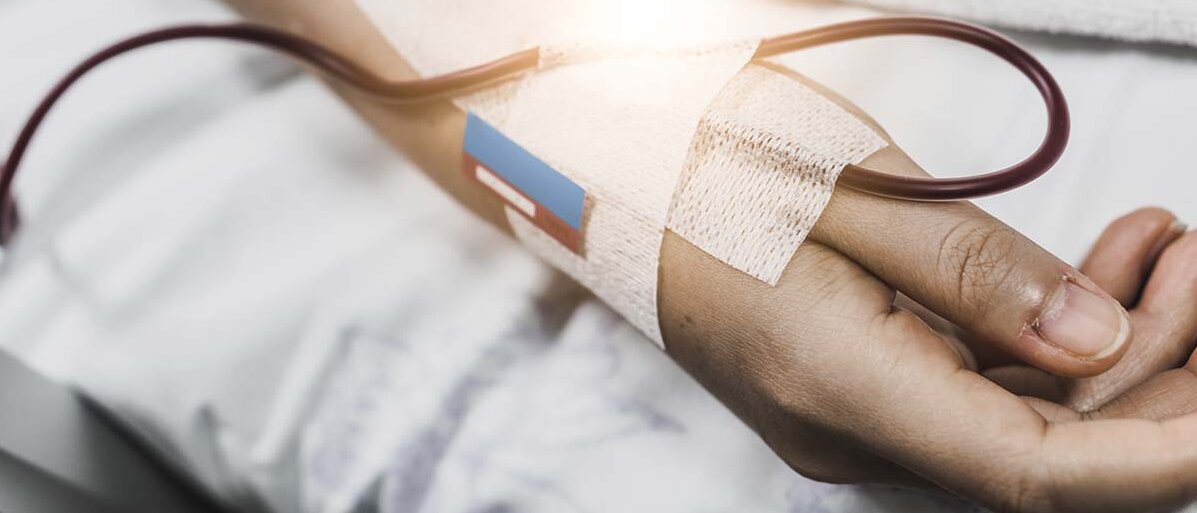 Eine Hand liegt auf weißer Bettwäsche, vermutlich auf einem Krankenhausbett. Ein Infusionsschlauch ist mit weißem Pflaster an der Hand befestigt.