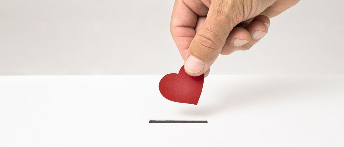 Eine Hand wirft ein Herz aus Papier in den Briefschlitz einer weißen Box.