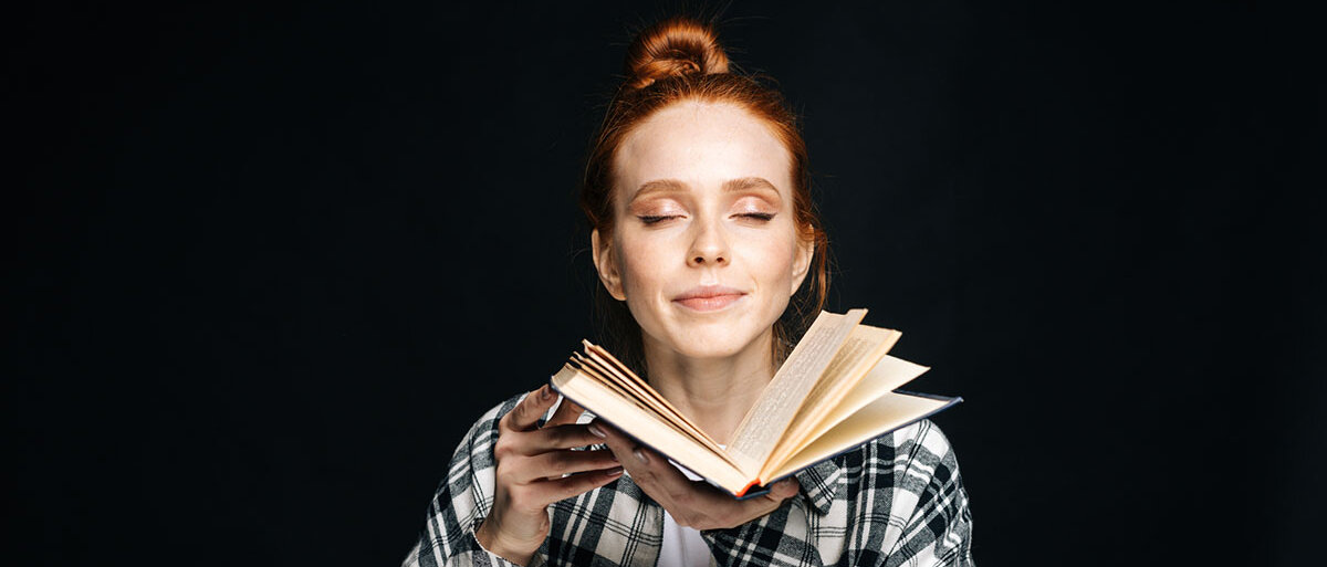 Eine junge Frau hält ein aufgeschlagenes Buch in den Händen, riecht daran und lächelt.