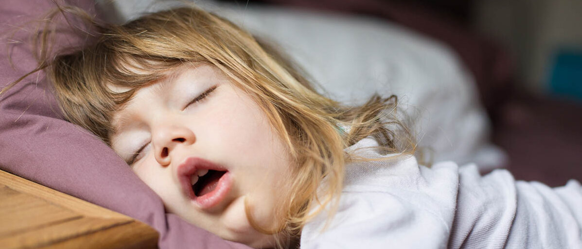 Melatonin in Gummibärchen: oft zu hohe Dosierung Ein kleines Mädchen schläft in Bauchlage. Ein Arm hängt aus dem Bett und der Mund steht offen.