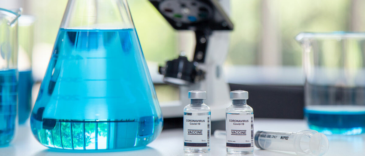 Auf einem Labortisch stehen ein Mikroskop, Erlenmeyerkolben und Bechergläser mit einer blauen Lösung und zwei Vials mit der Aufschrift "Coronavirus COVID-19 Vaccine".