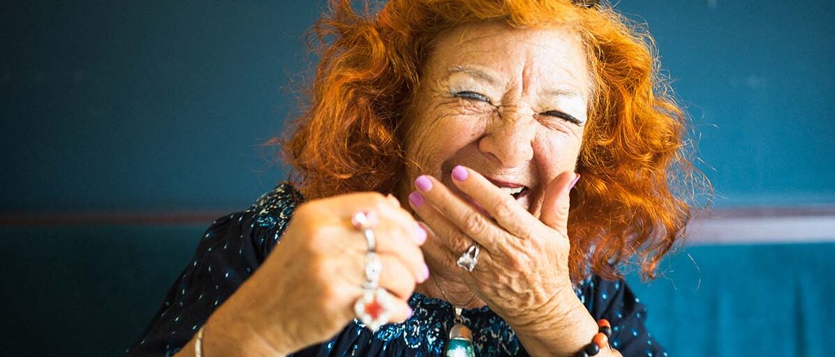 Porträt einer älteren, rothaarigen Dame, die herzhaft in die Kamera lacht.