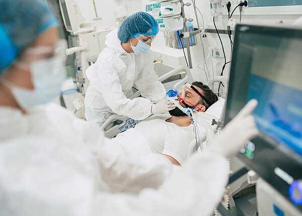 Das Guillain-Barré-Syndrom tritt häufig nach Infektionen mit bestimmten Keimen auf. Auch ein Zusammenhang mit Vektorimpfstoffen gegen COVID-19 ist bekannt. Ein Mann liegt im Krankenhaus auf der Intensivstation. Um sein Bett stehen zwei Pflegekräfte. Er wird beatmet.