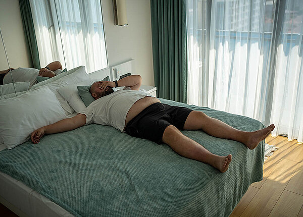 Ein Mann liegt bewegungslos im Bett und hält sich erschöpft die Hand ans Gesicht.