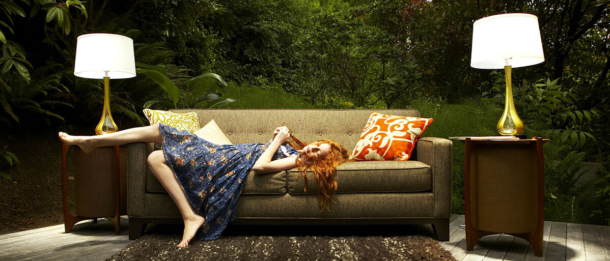 Eine Frau liegt auf einem Sofa, spielt mit ihren Haaren und hat ein Bein über die Lehne gelegt. Um das Sofa sind Beistelltische, Lampen und ein Teppich angeordnet und das Sofa steht auf einem Holzboden; das Ganze befindet sich aber nicht in einem Zimmer, sondern im Wald.