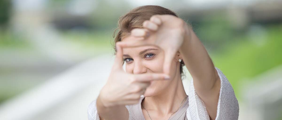 Eine Frau bildet mit den Händen ein Rechteck wie für einen Kameraausschnitt vor ihrem Gesicht. Durch dieses Rechteck schauen wir ihr in die Augen.