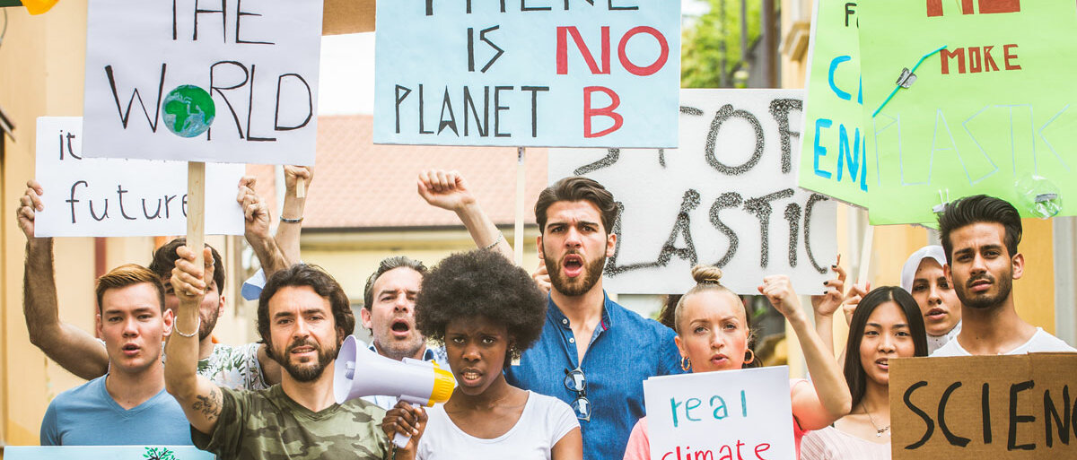 Mehrere Personen verschiedenen Alters und verschiedener Ethnien demonstrieren gemeinsam gegen den Klimawandel. Sie halten Schilder aus Pappe hoch, auf denen beispielsweise "There is no planet B" steht.
