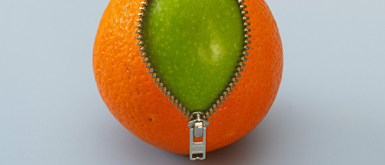 Eine Orange trägt einen Reißverschluss, der leicht geöffnet ist. DArunter kommt ein grüner Apfel zum Vorschein.