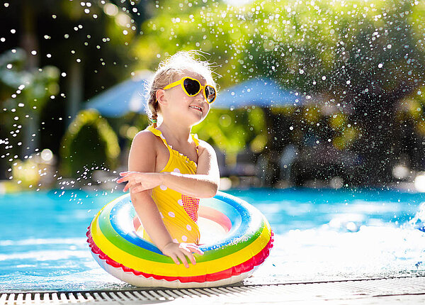 Mädchen trägt eine gelbe Herz-Sonnenbrille, einen gelben Badeanzug und einen Regenbogen-Schwimmreif. Sie steht am Schwimmbeckenrand und spritzt mit Wasser.