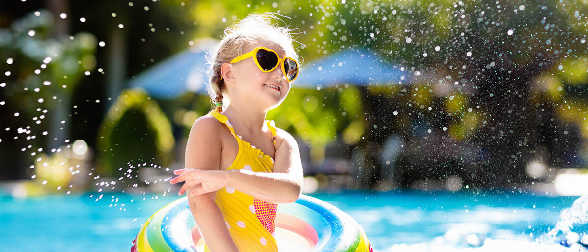 Mädchen trägt eine gelbe Herz-Sonnenbrille, einen gelben Badeanzug und einen Regenbogen-Schwimmreif. Sie steht am Schwimmbeckenrand und spritzt mit Wasser.
