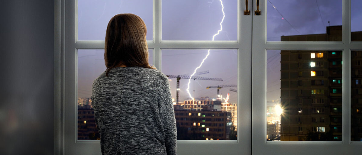 Junge Frau sitzt vor geschlossenem Fenster und schaut Gewitter zu