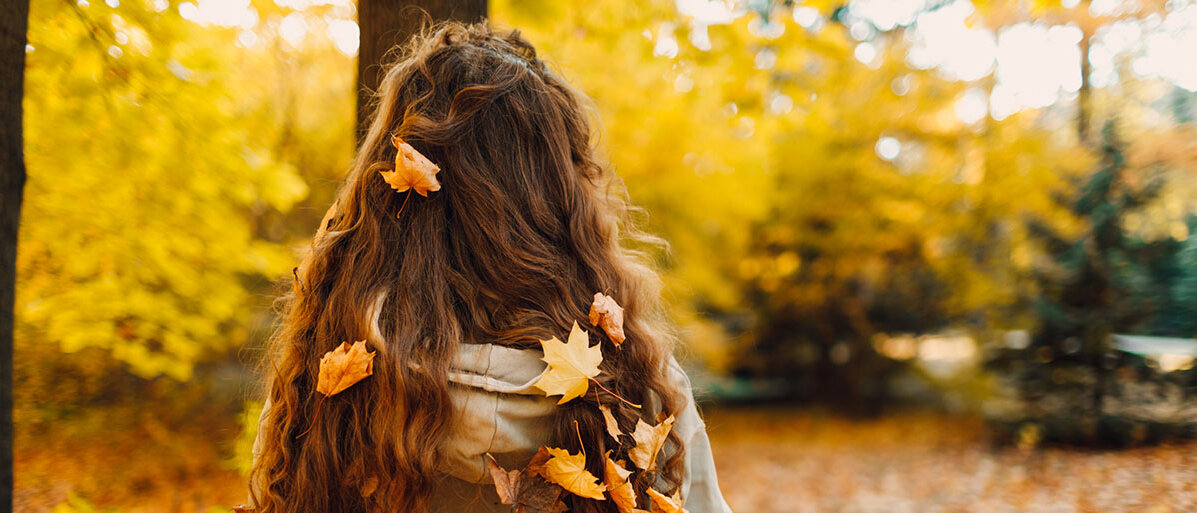 Eine Frau steht im Herbstwald. In ihren Haaren haben sich viele gelbe Blätter verfangen.