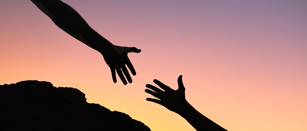 Zwei Hände nähern sich an. Im Hintergrund Sonnenuntergang. 