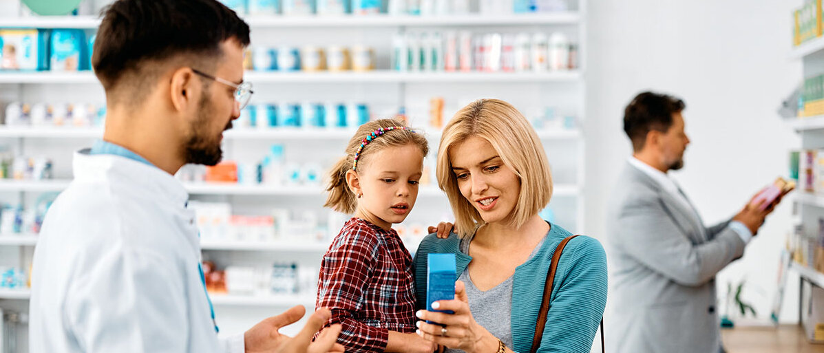 Ein Apotheker oder PTA berät eine Mutter, die ihre Tochter im Vorschulalter auf dem Arm hat. Die Mutter hält eine blaue Arzneimittelpackung in der Hand.