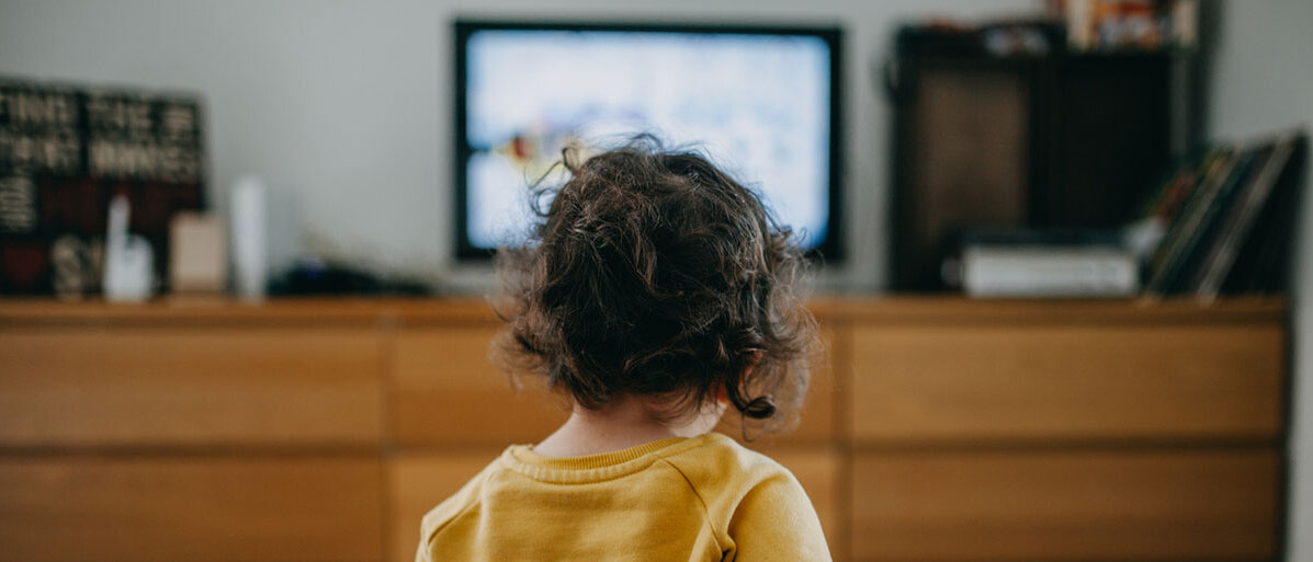 Kleines Maedchen mit gelbem Pullvoer und dunklen Haaren sitzt vor dem Fernseher
