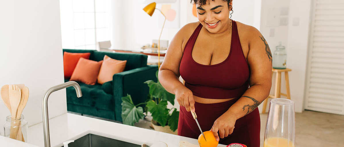 Eine Frau in Sportkleidung steht am Küchentresen und schneidet Obst auf für einen Smoothie.