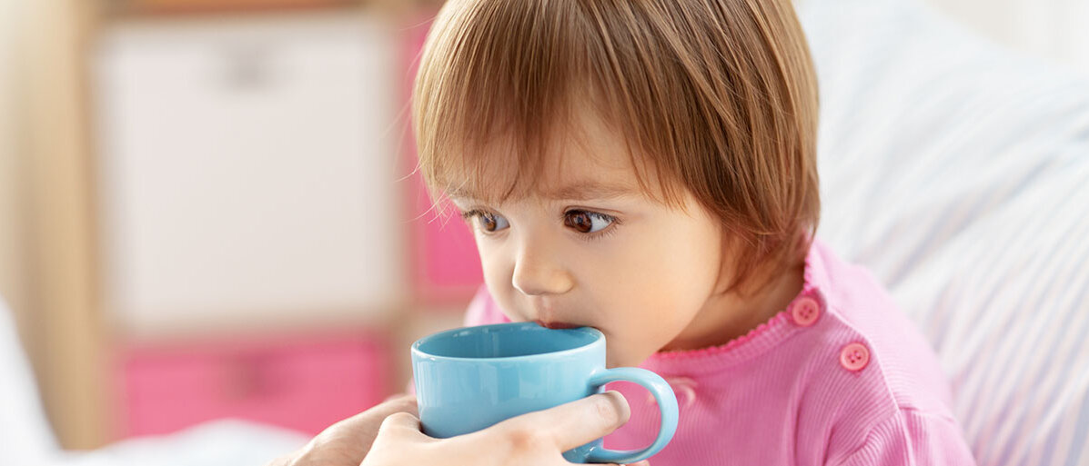 Kleines Maedchen mit rosa Pullover bekommt eine blaue Tasse mit Tee zum trinken an den Mund gehalten