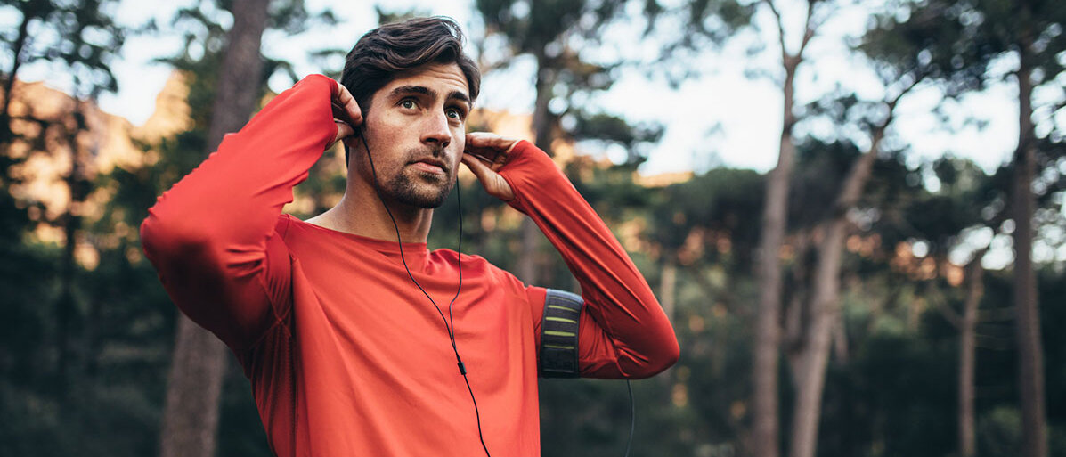 Man traegt rotes Trainingshirt und steht im Wald, hat Kopfhörer im Ohr