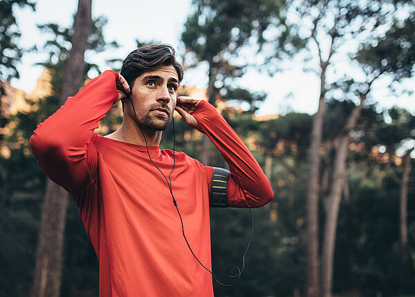 Man traegt rotes Trainingshirt und steht im Wald, hat Kopfhörer im Ohr
