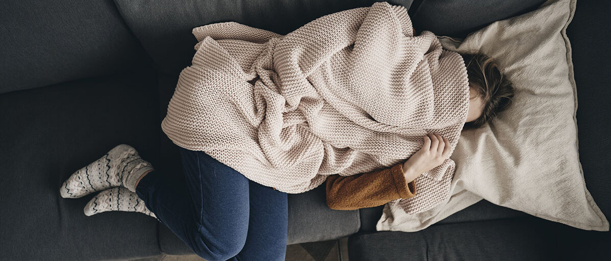 Eine junge Frau liegt mit angezogenen Beinen auf dem Sofa und hat sich die Decke über den Kopf gezogen.