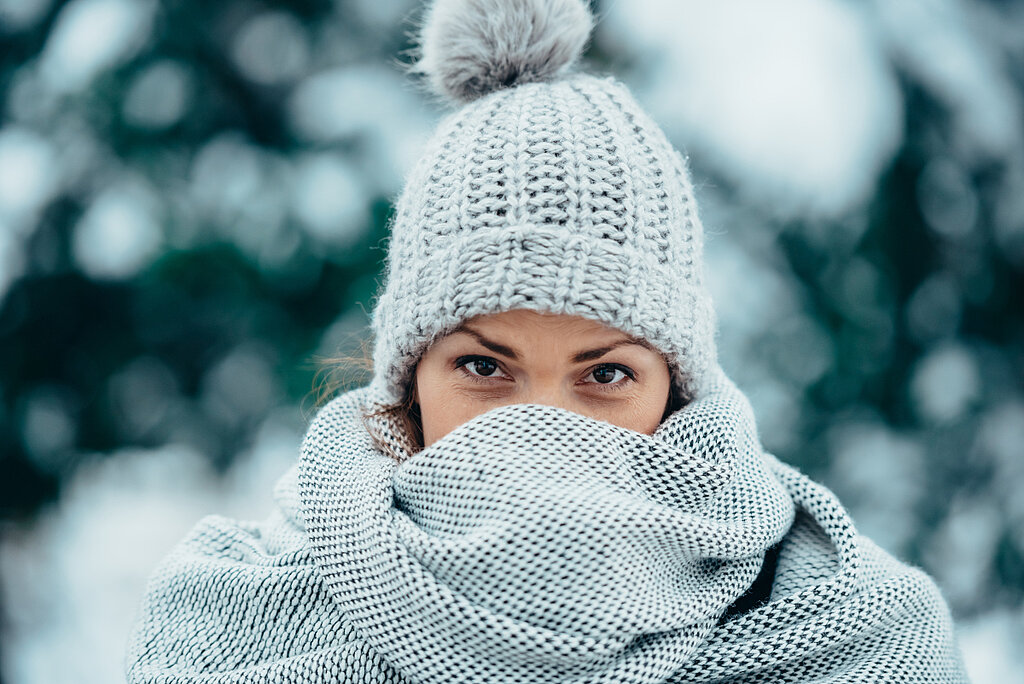 Es hat geschneit. Eine Frau steht im Freien, sie trägt eine Mütze und hat sich ihren Schal bis über die Nase gezogen.