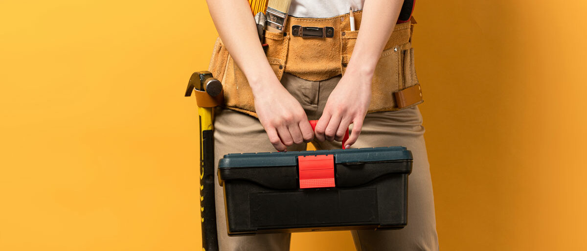 Eine Handwerkerin trägt einen Werkzeuggürtel und hält einen Werkzeugkasten.