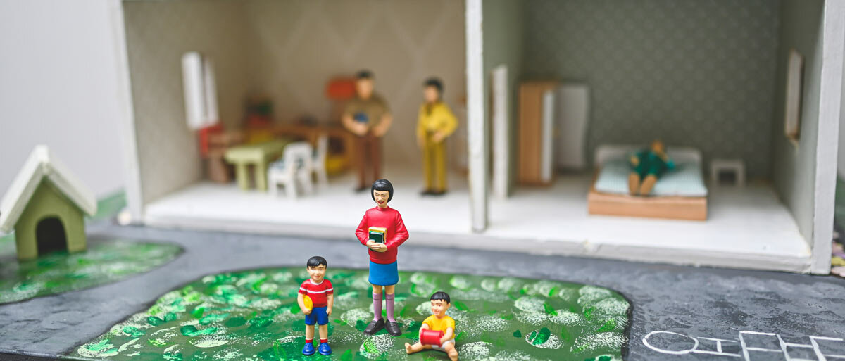 Eine Puppenfamilie im Puppenhaus: Die Mutter und zwei Söhne spielen im Garten, zwei Männer stehen in der Küche, eine Person schläft im Schlafzimmer.