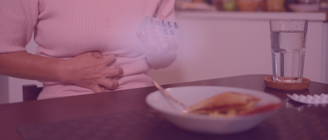 Eine Frau hat einen halb aufgegessenen Teller vor sich stehen, greift sich verkrampft an den Bauch und hält einen angebrochenen Blister mit Kapseln in der Hand. Der Fokus liegt auf dem Medikament.