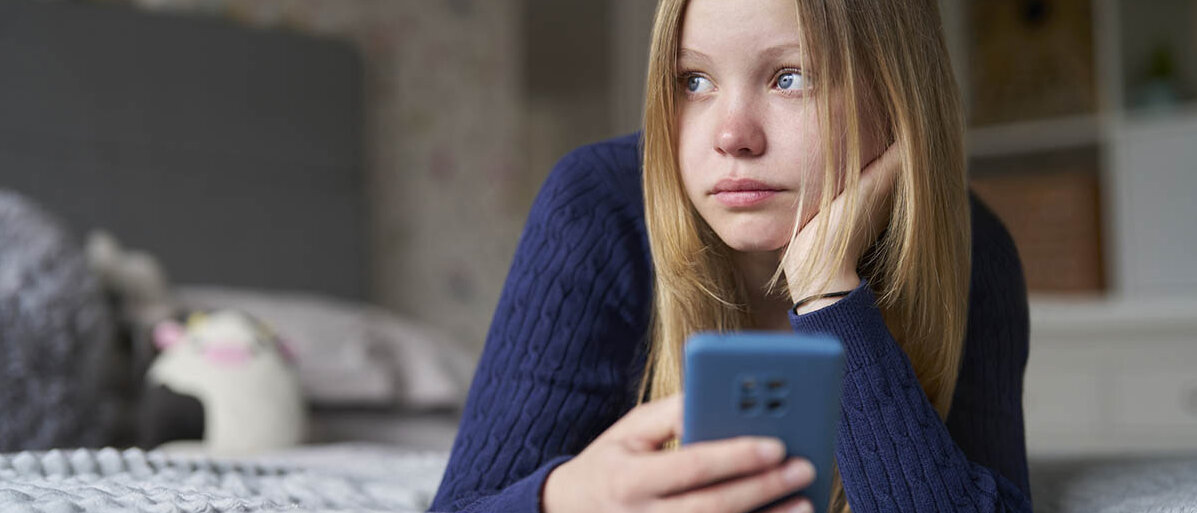 Gesunde Handynutzen bei Jugendlichen: Tipps für den Smartphone-Konsum Ein Mädchen liegt auf dem Bauch auf ihrem Bett, hat den Arm aufgestützt und ihr Smartphone in der Hand und schaut aus dem Fenster.
