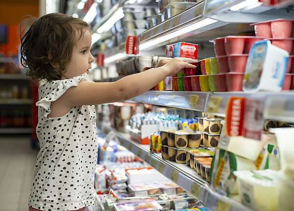 Kleines Mädchen steht im Supermarkt vor einem Regal mit Joghurt und greift nach einer Sorte