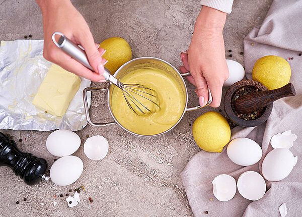 Herstellung traditioneller Sauce Hollandaise in einem Topf; daneben weiße Eier, Zitronen, Butter