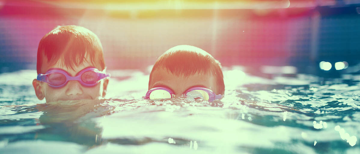 Zwei Jungen im Freibad: Beide haben das Gesicht halb über, halb unter Wasser und tragen eine Schwimmbrille.