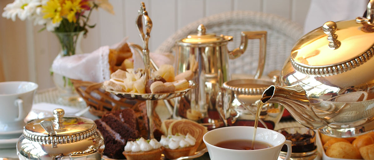 Silberkanne schenkt Tee in Tasse ein, nebendran eine Etagere, Zuckerdose und weitere Elemente einer klassischen englischen Tea Time