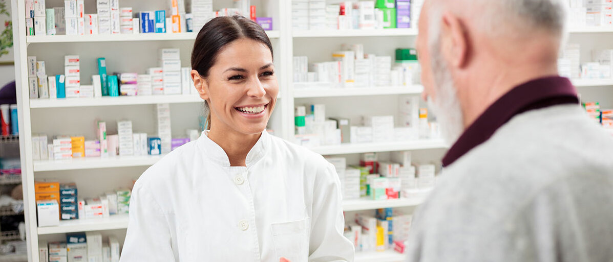 Eine Apothekerin oder PTA reicht einem Kunden ein Arzneimittelpäckchen über den HV-Tisch.
