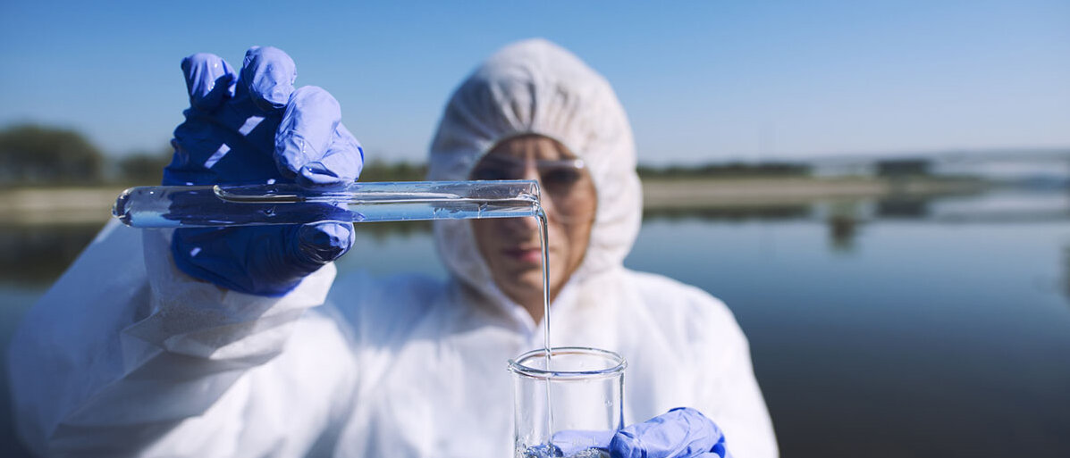 Eine Person im weißen Schutzanzug steht vor einem See und gießt eine Wasserprobe aus einem Reagenzglas in ein Becherglas.