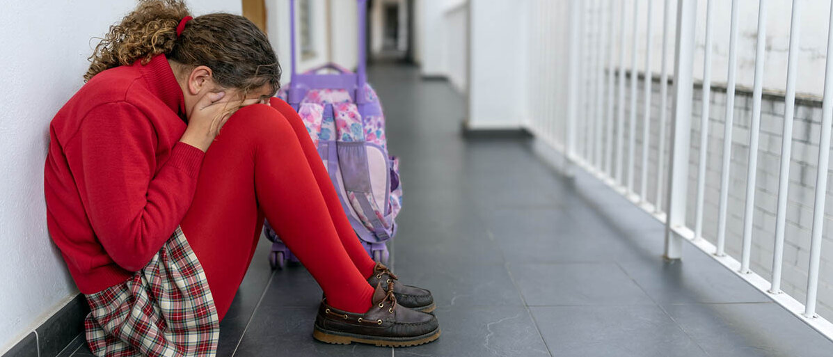 Regelschmerzen von Teenagern vorbeugen und sie behandeln: Welche Medikamente eignen sich bei Jugendlichen mit Periodenschmerzen? Eine Schülerin sitzt allein im Flur der Schule, Beine angezogen, Gesicht in den Händen verborgen.