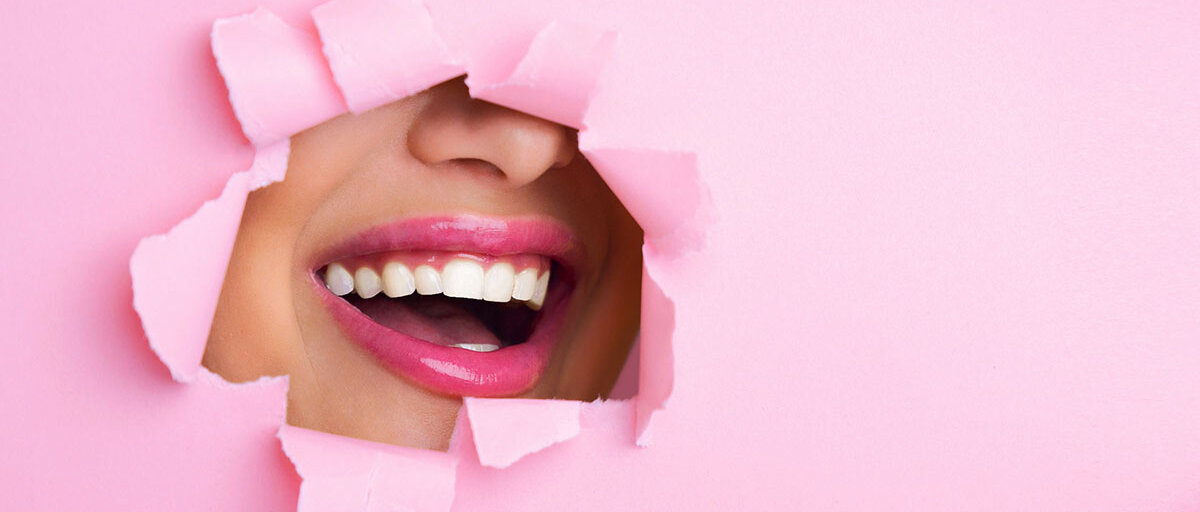 Eine rosa Papierwand ist aufgebrochen, dahinter sieht man einen lachenden Frauenmund mit pink geschminkten Lippen.